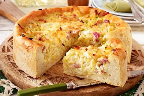 Ein goldbrauner Zwiebelkuchen mit Speck, ein köstliches zwiebelkuchen originalrezept, auf einem Holzbrett mit einem Messer.