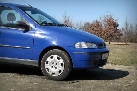 Błękitny Fiat Albea stoi na polnej drodze, w tle drzewa i trawa.