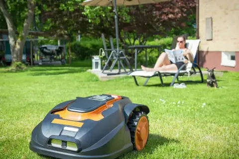 Automatyczna kosiarka do jezdzenia pracuje na trawniku, podczas gdy kobieta odpoczywa na leżaku.
