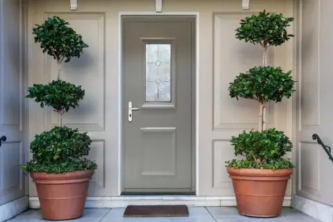 Une porte d'entrée grise avec une fenêtre décorative, encadrée par deux grands pots de plantes taillées. Idéal pour savoir comment changer une porte d'entrée.