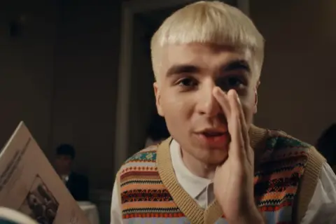 Młody mężczyzna z blond włosami, w kamizelce w kolorowe wzory, trzyma w ręku kartkę z tekstem, jakby czytał papuga.