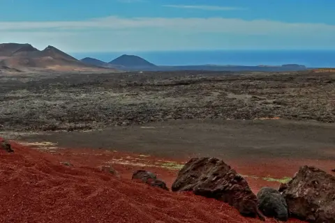 Krajobraz w parku narodowym Timanfaya: czerwone wzgórza, pola lawy i góry na tle oceanu.
