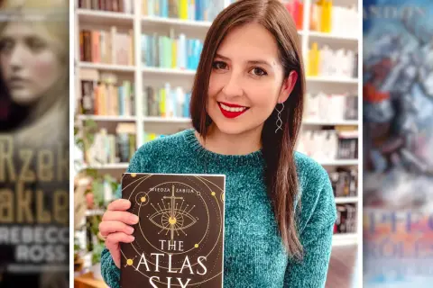 Kobieta w swetrze trzyma książkę "The Atlas Six", otoczona okładkami najlepszych serii fantasy.