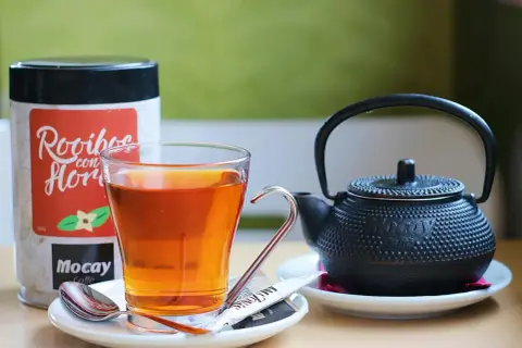 Szklanka z bursztynowym naparem, obok czajniczek i puszka z napisem "Rooibos con Flor". Czas na chwilę relaksu z rooibos.
