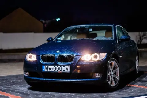 Ciemnoniebieskie bmw 325i z kroplami deszczu na karoserii, zaparkowane nocą na mokrym parkingu.