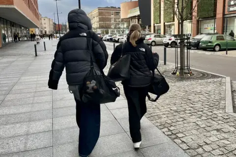 Dwie osoby w czarnych kurtkach puchowych idą chodnikiem, niosąc torby. Proste, miejskie kurtka puchowa stylizacje.