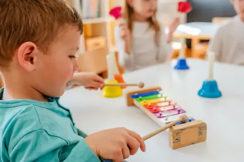Chłopiec aktywnie w żłobku gra na cymbałkach, obok inne dzieci bawią się dzwonkami.