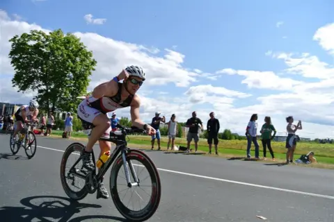 Triathlonista w kasku i okularach ściga się na rowerze, a widzowie dopingują go na poboczu. To część kalendarza triathlonowego.