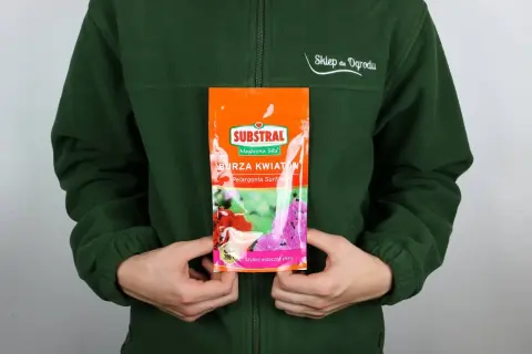 Opakowanie nawozu Substral Burza Kwiatów z pelargoniami, trzymane przez osobę w zielonej bluzie.