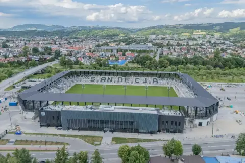 Nowoczesny stadion Sandecji z zieloną murawą i trybunami, otoczony miastem i wzgórzami.