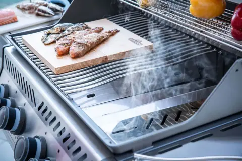 Fisch auf einem Holzbrett auf einem Grill, der zeigt, was ein Smoker Grill kann: Rauch steigt auf und veredelt das Gargut.