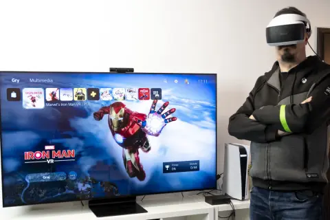 Mężczyzna w goglach VR obok telewizora z grą Iron Man VR.