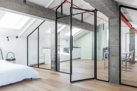 Moderno recupero sottotetto con pareti in vetro, travi a vista in cemento e parquet chiaro. Ambiente luminoso con cucina e zona living.