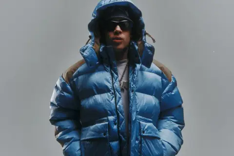 Giovane con piumino blu acceso, cappuccio alzato, occhiali da sole e una collana con ciondolo. Lo sfondo è un grigio chiaro, quasi low red.