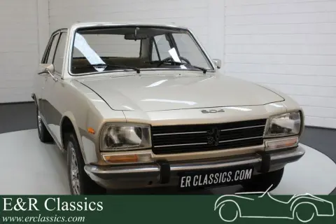 En ljusbeige Peugeot 504 står i ett garage. Bilen har en klassisk design med kromade detaljer.