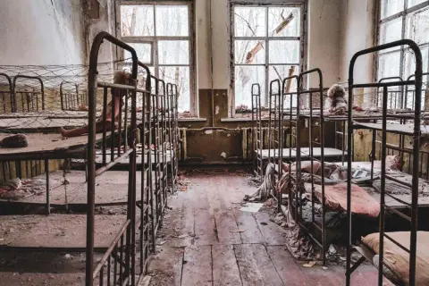 Opuszczony pokój z rzędami zardzewiałych łóżek piętrowych, zniszczonymi oknami i śmieciami na podłodze.