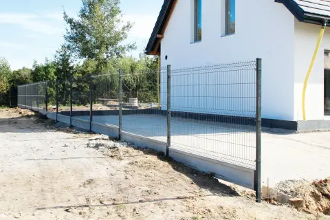Trwa budowa ogrodzenia wokół nowoczesnego domu. Metalowa siatka na betonowym fundamencie otacza posesję.