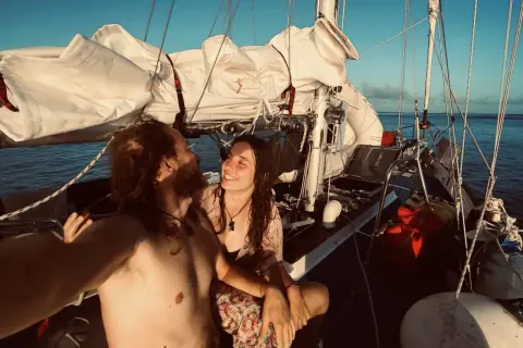 Coppia felice in barca a vela al tramonto, con vele bianche spiegate e mare calmo.