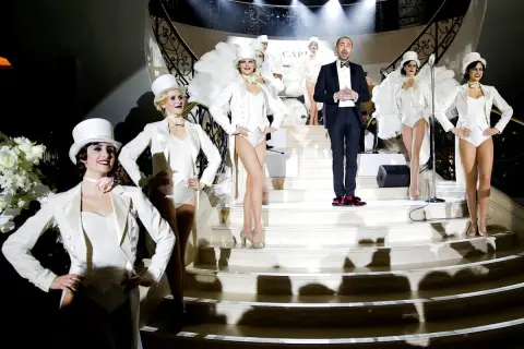 Bühnenauftritt mit Tänzerinnen in weißen Outfits und einem Mann im schwarzen Anzug mit **white tie**.