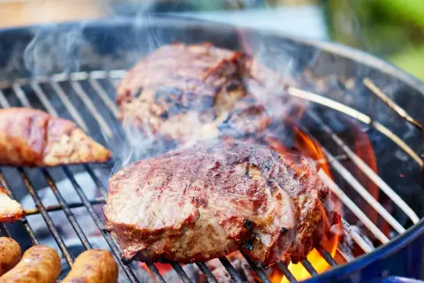 Saftiges Fleisch zum Grillen zischt auf dem Rost. Würstchen und Steaks brutzeln über heißen Kohlen, umgeben von Rauch und Flammen.