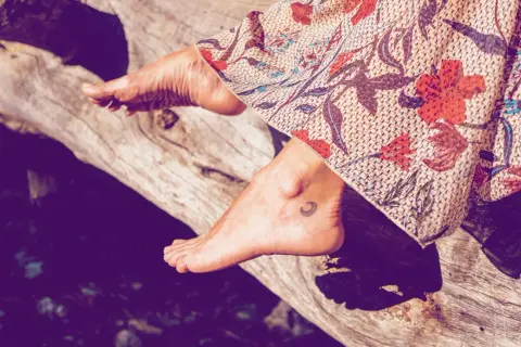 Ein Fuß mit einem kleinen Mond-Tattoo ruht auf einem Baumstamm, bedeckt von einem gemusterten Stoff.