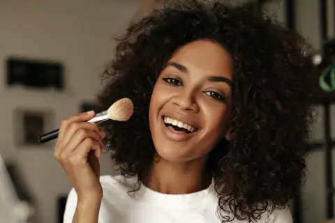 Donna sorridente con capelli ricci applica cipria con un pennello, mostrando come mettere la cipria senza pennello.