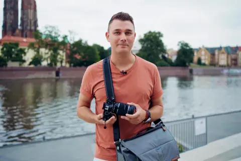 Mężczyzna z aparatem w rękach, w plenerze nad rzeką, z widokiem na zabytkowe budynki i katedrę.