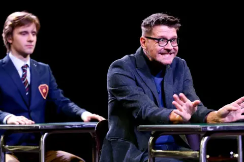 Dwóch aktorów na scenie podczas spektaklu "Stowarzyszenie Umarłych Poetów". Młody mężczyzna w garniturze i starszy z okularami, obaj siedzą przy stołach.