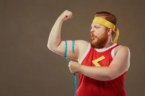 Mężczyzna typu endomorfik z wąsami i brodą, w żółtej opasce i czerwonej koszulce, mierzy obwód bicepsa.