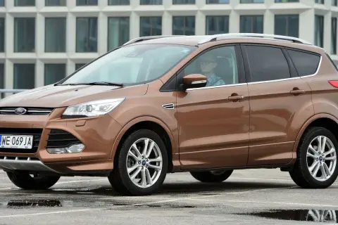 Brązowy Ford Kuga 2 z kierowcą w okularach i kratę, zaparkowany na mokrym parkingu przed nowoczesnym budynkiem.