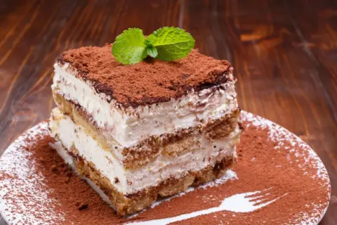 Ein Stück cremiges Tiramisu ohne Ei, bestäubt mit Kakao und garniert mit Minze, auf einem Teller mit Kakaopulver-Gabelabdruck.