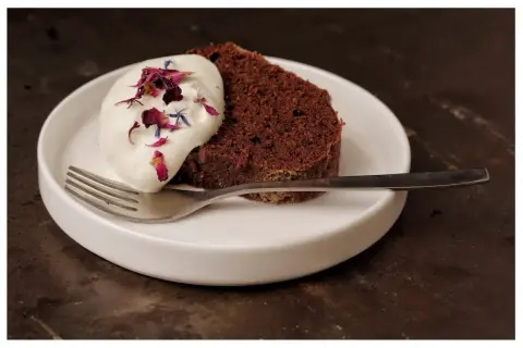 En bit saftig sockerkaka choklad med vispgrädde och blomblad på en vit tallrik med en gaffel.