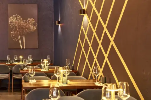 Stilvolles Ambiente im Lovis Restaurant: gedeckte Tische mit Gläsern und Kerzen, dunkle Wände mit goldenem Muster und ein Kunstwerk mit getrockneten Blumen.