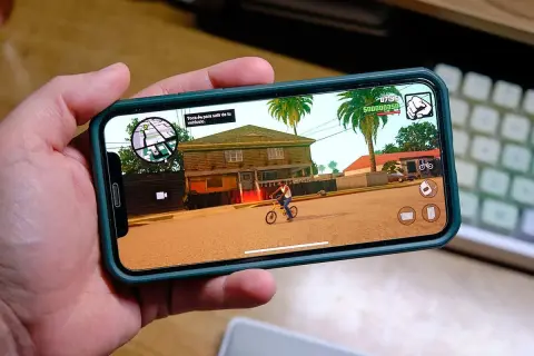Ręka trzyma smartfon z grą RPG na telefon. Na ekranie widać postać na rowerze przed domem, palmy i ikony sterowania.