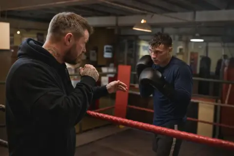 A boxer trains with his coach in a boxing ring. The coach, with tattoos on his arms and neck, demonstrates a move. The boxer, wearing gloves, listens intently. This is a scene of dedication and the pursuit of the perfect box weight.