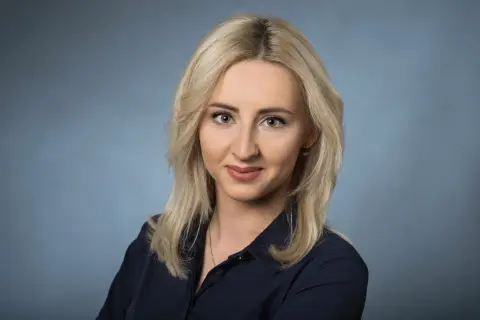 Portret uśmiechniętej blondynki w granatowej koszuli na niebieskim tle.