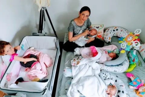 Mama karmi jedno z czworaczków, podczas gdy starsza siostra opiekuje się drugim dzieckiem w łóżeczku.