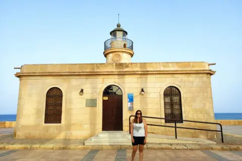 Mujer sonriente posa frente al Faro de Roquetas de Mar, con el mar azul de fondo.