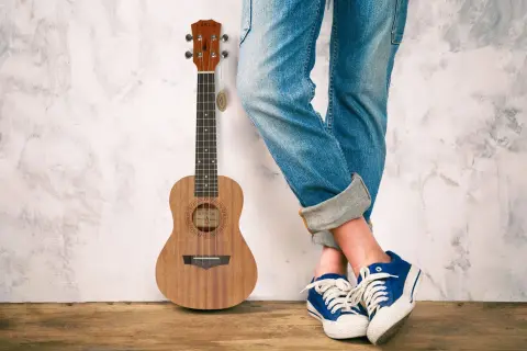 Drewniane ukulele stoi obok osoby w jeansach i niebieskich trampkach. To małe, czterostrunowe ukulele.