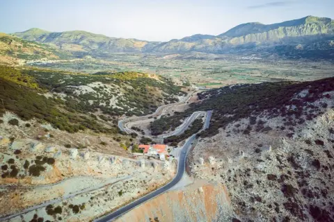 Vue aérienne d'une route sinueuse traversant un paysage montagneux verdoyant, menant à un village sur le plateau de Lassithi. Des ruines de moulins à vent parsèment la colline.