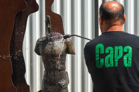 Hombre con camiseta negra con letras verdes "Capa" limpia una estatua de bronce con agua a presión.