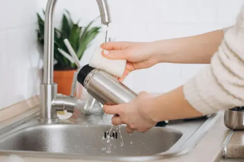 Hände reinigen eine Edelstahl-Thermoskanne unter fließendem Wasser im Spülbecken.