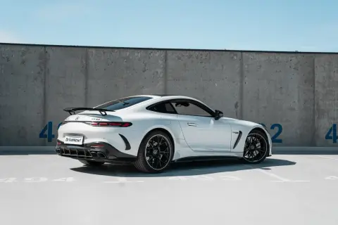 Biały Mercedes GT 63 AMG z czarnym spoilerem i felgami, zaparkowany pod betonową ścianą z niebieskimi cyframi.