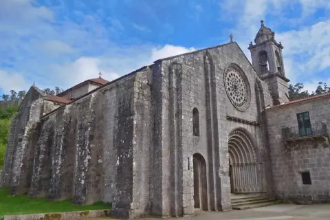 El monasterio de Armenteira, con su imponente fachada de piedra y su rosetón, se alza bajo un cielo azul con nubes.