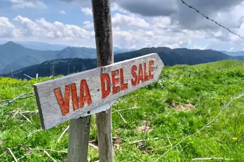 Cartello in legno indica il **via del sale cammino**. Panorami montani verdi e cielo azzurro con nuvole bianche.