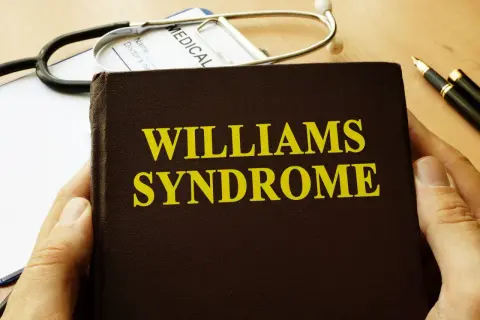 Dłoń trzyma książkę z napisem "Williams Syndrome". Obok leży stetoskop i długopis.