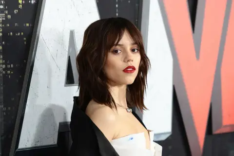 Jenna Ortega mit mittellangem Bob mit Pony und rotem Lippenstift, posiert vor einem großen Logo.