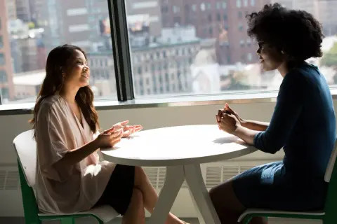 Dos mujeres conversan animadamente en una oficina, con una vista urbana de fondo. Una de ellas gesticula mientras habla, demostrando un nivel de **speaking B2** en su comunicación.