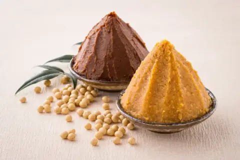 Dwa stożki pasty miso, jasnej i ciemnej, w miseczkach, obok ziaren soi i liści. Co to miso?