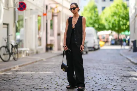 Eine Frau trägt ein festliches Oberteil zu schwarzer Hose, Sonnenbrille und einer kleinen schwarzen Tasche.